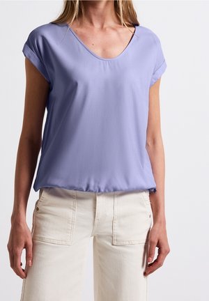 Kvinde iført lys lilla kortærmet bluse med opviklede manchetter og lyse beige bukser med store forlommer.