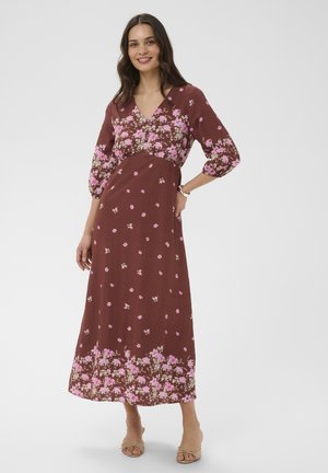 Femme debout souriante portant une robe longue marron à motifs floraux roses, manches trois-quarts, et des talons beiges ouverts au bout, sur fond blanc.