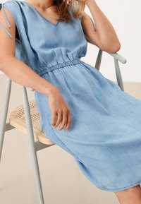 Ljusblå denimklänning med V-ringning, samlad midja och knytärmar. Slät textur och knälång design, sittande på en stol.