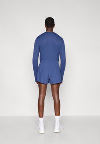 Μπλε μπλούζα με μακριά μανίκια και ταιριαστά σορτς από ελαφρύ ύφασμα, με ελαστική ζώνη, σε συνδυασμό με λευκά αθλητικά παπούτσια και κάλτσες, από πίσω.