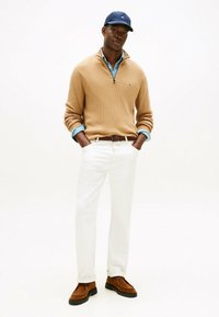 Uomo che indossa un maglione color sabbia con cerniera a mezza altezza sopra una camicia blu, pantaloni bianchi, cintura marrone, scarpe in camoscio marrone e un berretto blu navy, in piedi con le mani nelle tasche.