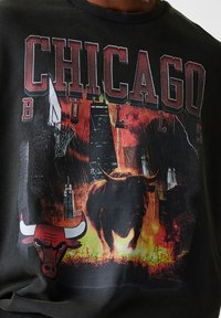 Sweatshirt noir avec un graphisme d'un taureau en charge, accompagné du texte "CHICAGO BULLS" et d'un arrière-plan de skyline de la ville dans des teintes rouges et jaunes.