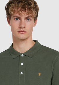 Olivgrünes Poloshirt aus strukturiertem Stoff, mit Knopfleiste, kurzen Ärmeln und einem gestickten orangen Logo auf der Brust.