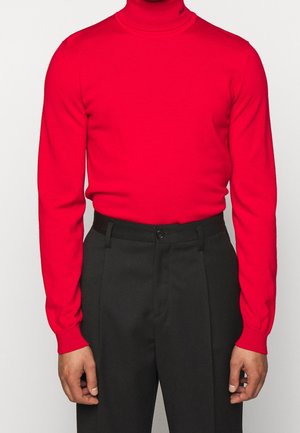 Red ribbed turtleneck sweater with long sleeves, paired with high-waisted black trousers featuring a front button closure.