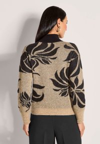 Frau mit langen dunklen Haaren, die einen beigen Pullover mit großen schwarzen Blumenmustern und schwarze Hosen trägt, mit dem Rücken zu einer einfarbigen Hintergrund.