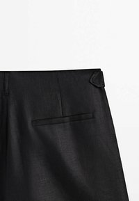 Pantalon noir sur mesure avec une poche passepoilée horizontale à l'arrière et un détail de patte de ceinture sur fond blanc.