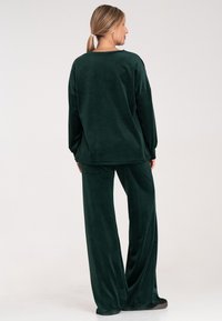 Zestaw dresowy z ciemnozielonego weluru składa się z luźnej bluzy z długim rękawem i spodni o szerokiej nogawce, prezentując gładką teksturę i swobodny styl.