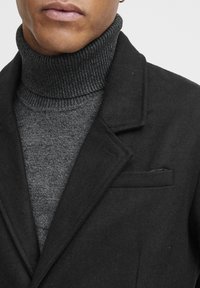 Abrigo de lana negra con solapa recortada, superpuesto a un cuello de tortuga de canalé gris oscuro. Textura suave y diseño estructurado. Detalle de bolsillo visible.