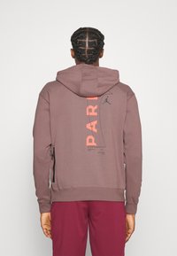 Sweat à capuche mauve avec un motif graphique "PART" corail dans le dos, comprenant un logo noir et des fermetures éclair latérales en détail.