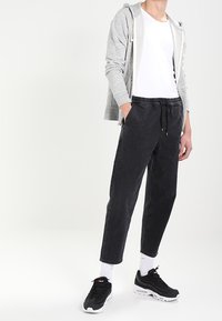 Person som bär en grå zip-up hoodie, vit t-shirt, svarta dragsko-byxor, vita strumpor och svarta träningsskor, stående med händerna i fickorna.