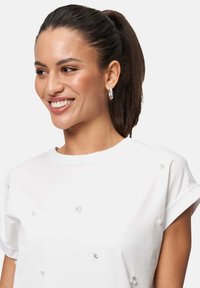 T-shirt blanc à manches courtes avec des accents de perles décoratives, encolure ronde et coupe décontractée. Modèle présenté avec des cheveux raides et foncés et des boucles d'oreilles créoles.