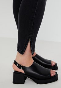 Black heeled sandals with open toe and ankle strap, featuring a smooth leather texture and a chunky square heel. Jeans with zippers at the ankles.