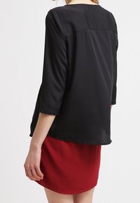 Blusa nera semi-trasparente con maniche a tre quarti e orlo arrotondato, abbinata a una gonna rossa. Tessuto liscio e design semplice.