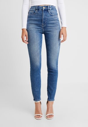 Kvinde iført højtaljede blå skinny jeans og hvid langærmet top, stående i hvide åbne hæle på ensfarvet baggrund.