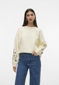 Sudadera de algodón crema con bordado floral en las mangas, escote redondo y ajuste relajado, combinada con jeans azules de cintura alta.