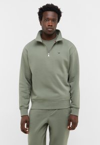 Mężczyzna w oliwkowej, rozpinanej bluzie i dopasowanych spodniach, stojący przed gładkim białym tłem z neutralnym wyrazem twarzy.