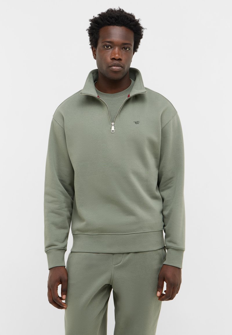 Mężczyzna w oliwkowej, rozpinanej bluzie i dopasowanych spodniach, stojący przed gładkim białym tłem z neutralnym wyrazem twarzy.