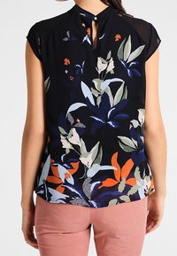 Blouse fleurie bleu marine foncé avec manches courtes et détail dos à ouverture en goutte d'eau. Présente des motifs botaniques verts, orange et blancs. Texture lisse.