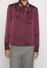 Blusa di raso bordeaux con collo a fiocco, maniche lunghe con polsini a bottone e una texture liscia. Abbinata a pantaloni con motivo.