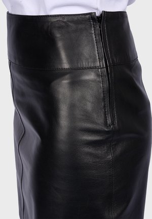 Jupe en cuir noire taille haute avec détail de fermeture éclair latérale portée avec une chemise blanche rentrée sur fond neutre.