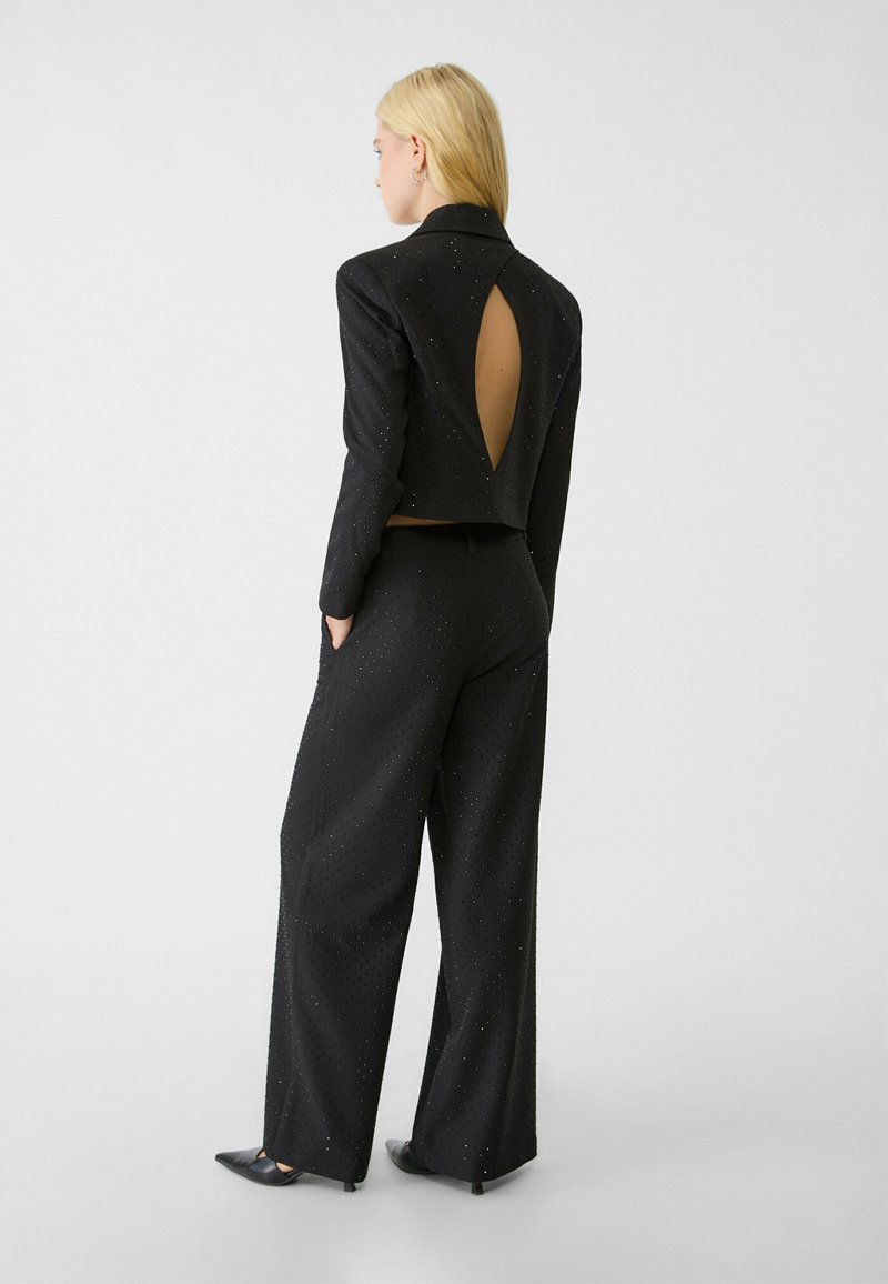 Conjunto de dos piezas en negro que presenta una chaqueta corta con un corte en la parte posterior y detalles brillantes, combinada con pantalones de pierna ancha hechos de tela texturizada.
