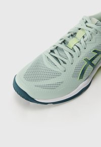Chaussure de sport vert clair avec tige en maille, détails texturés et semelle bleu foncé. Elle est dotée de lacets et d'un logo bien visible dans une teinte plus foncée.