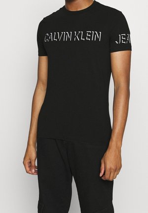 Person trägt ein eng anliegendes schwarzes Calvin Klein T-Shirt mit weißem Logo auf der Brust und teilweise weißem Text auf dem Ärmel.