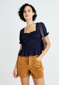 Navyblauw peplumblouse met geraffineerde textuur en kantafwerking, voorzien van kleine gouden accenten, gecombineerd met bruin suedeshorts.
