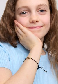 Czarna bransoletka z kordelkiem z złotym symbolem nieskończoności i koralikami. Kordelka ma regulowane zapięcia do dopasowania. Noszona na jasnoniebieskim rękawie.