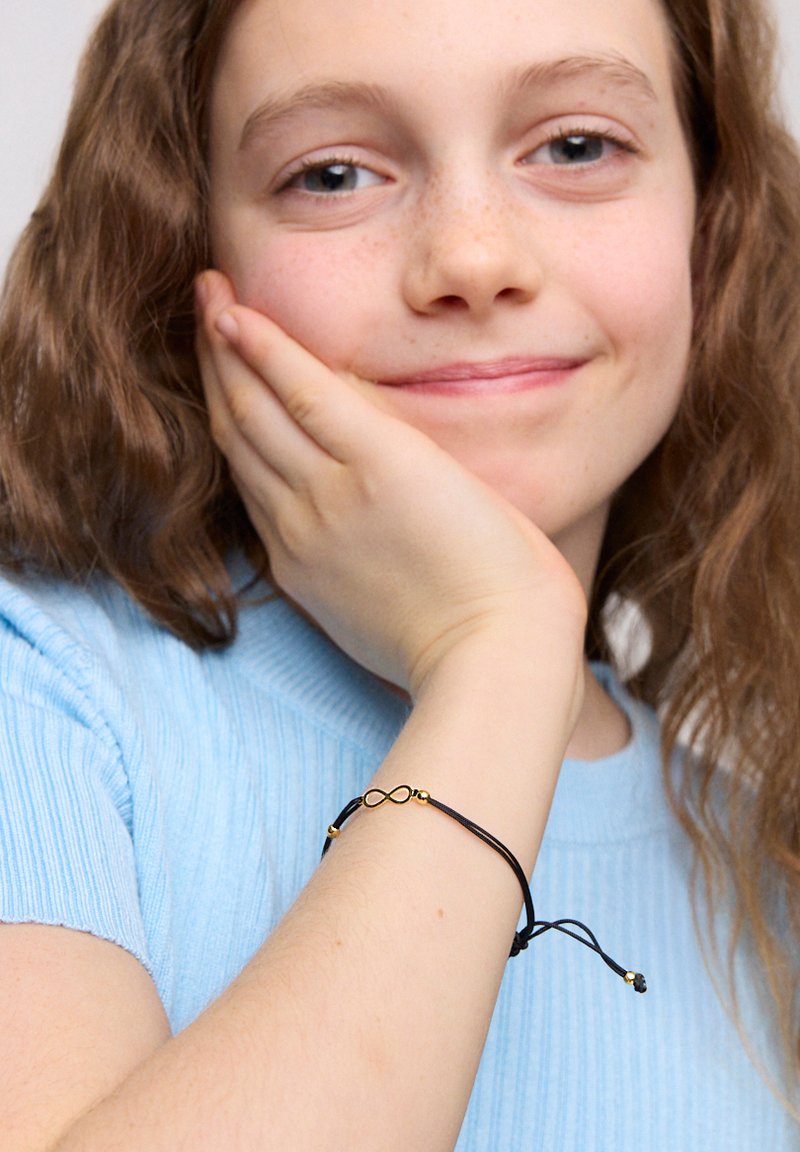Czarna bransoletka z kordelkiem z złotym symbolem nieskończoności i koralikami. Kordelka ma regulowane zapięcia do dopasowania. Noszona na jasnoniebieskim rękawie.