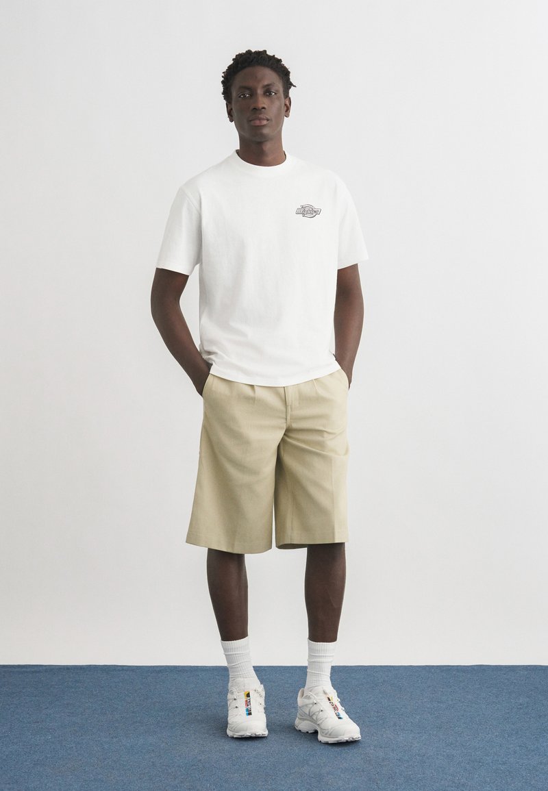 Homme debout, mains dans les poches, portant un short beige, un t-shirt blanc avec un petit logo, des chaussettes blanches et des baskets blanches sur un sol bleu devant un mur blanc.