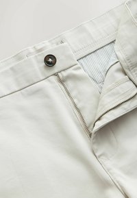 Gros plan de la ceinture d'un pantalon beige avec fermeture éclair ouverte, bouton fermé et doublure de poche rayée visible.