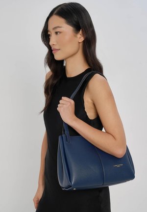 Femme en robe noire sans manches tenant un sac cabas bleu Lancaster sur l'épaule, tournée à gauche sur un fond uni.