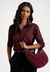 Borsa in suede bordeaux con forma curvata, dettagli in pelle e hardware in metallo. Il modello indossa una blusa e una cintura abbinate in bordeaux.