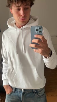Mladic u bijelom dukserici i plavim farmerkama snima selfie u ogledalu s pametnim telefonom u prostoriji s drvenim podom.