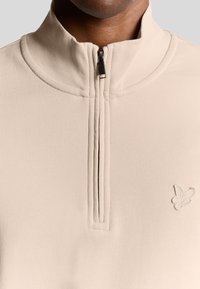 Maglione beige a zip verticale con tessuto texturizzato. Presenta un colletto alto e un piccolo logo di farfalla ricamato sul lato sinistro del petto.