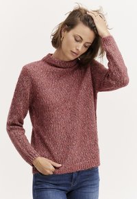 Krustotas džemperis silti rozā krāsā ar augstu apkakli, garām piedurknēm un teksturētu apdari, apvienots ar ziliem džinsiem.