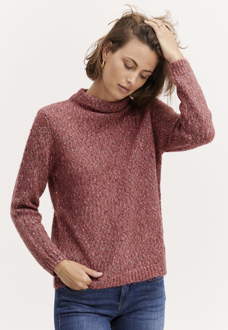 Krustotas džemperis silti rozā krāsā ar augstu apkakli, garām piedurknēm un teksturētu apdari, apvienots ar ziliem džinsiem.
