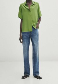 Chemise verte à col avec des manches courtes, associée à un jean droit bleu clair et des sandales noires. Le tissu semble lisse et léger.