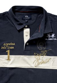 Marinblå pikétröja med vit rand, guldbroderi och knappar. Har texten "Argentina Polo Team" och en signatur, från La Martina. Storlek M.