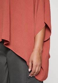 Rostorangefarbener, strukturierter Oberteil mit drapierten Ärmeln und ungleichmäßigem Saum. Getragen über dunkelgrauer Hose mit Seitentasche.