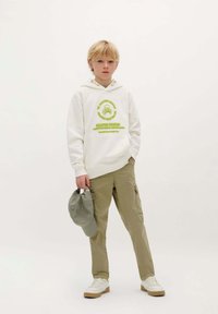 Witte hoodie met groene grafische print, khaki cargo pants met zijzakken, grijze baseballcap en witte sneakers met beige zolen.