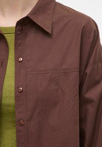 Bruine button-up shirt met een kraag, voorzien van een patchzak en textuurdetails. Gedragen over een groen, geribbeld T-shirt met lange mouwen.