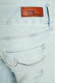 Jeans de mezclilla azul claro con un parche de cuero marrón que lleva "LTB". El bolsillo trasero tiene un bordado intrincado y un detalle de remache de metal plateado.