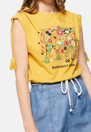 Frau trägt ein gelbes ärmelloses Oberteil mit floralem Elefantendesign und dem Text "Blooming Inside", kombiniert mit hellblauen Hosen und weißem Seilgürtel.