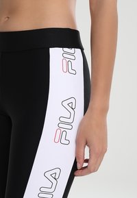 Närbild av en person som bär svarta leggings med ett vitt sidostråk prytt av upprepade FILA-logotyper i svart och röd längs låret.
