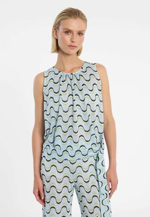 Frau trägt ärmelloses Oberteil und passende Hose mit gewelltem Muster in Schwarz, Grün und Hellblau auf weißem Hintergrund.