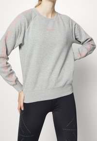 Felpa grigia con maniche lunghe, polsini e vita a costine. Presenta un marchio rosa lungo le maniche. Abbinata a leggings neri decorati con pois.
