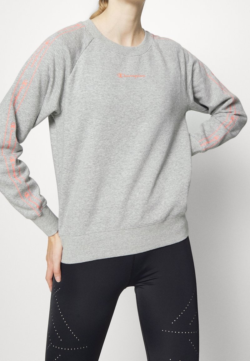 Felpa grigia con maniche lunghe, polsini e vita a costine. Presenta un marchio rosa lungo le maniche. Abbinata a leggings neri decorati con pois.