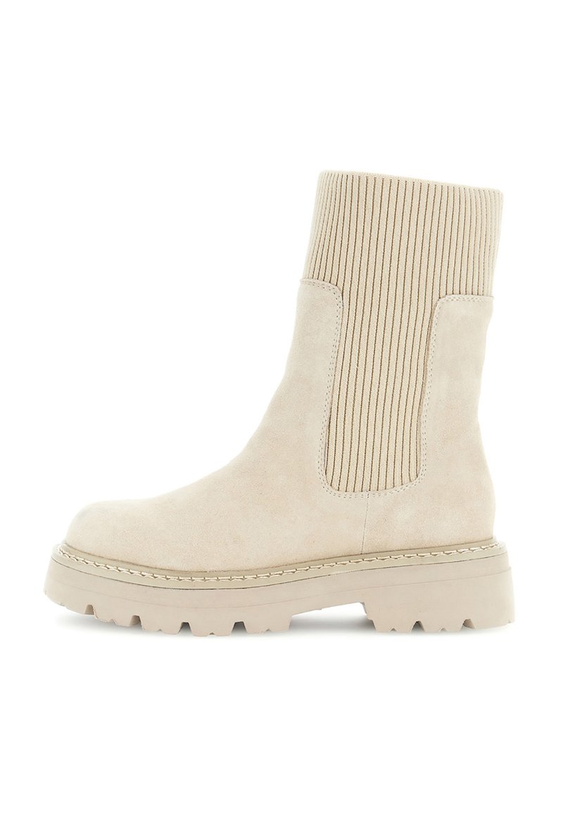 Beige ruskindsankelstøvle med ribbet strik elastisk sidepanel og tyk sål på en hvid baggrund.